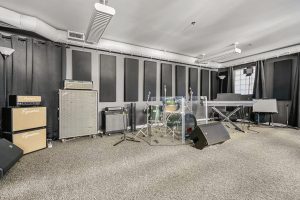 Studio room with drums, amps, keyboard, PA, and sound panels.